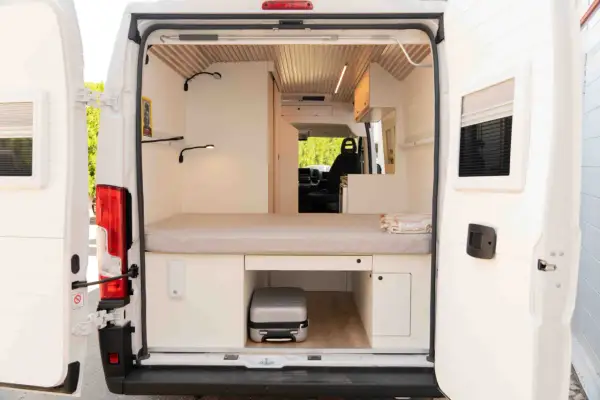 Interior de una camper con cama, almacenamiento y un pequeño equipaje. La altura camper permite aprovechar cada rincón.