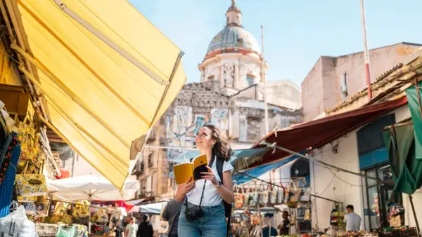 Turista sonriente con libro y cámara explora los vibrantes mercados de Palermo, con un domo histórico al fondo.