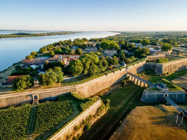 Vista aérea de la ciudadela de Blaye, con sus imponentes murallas de piedra, un puente arqueado y viñedos verdes junto a un río sereno.