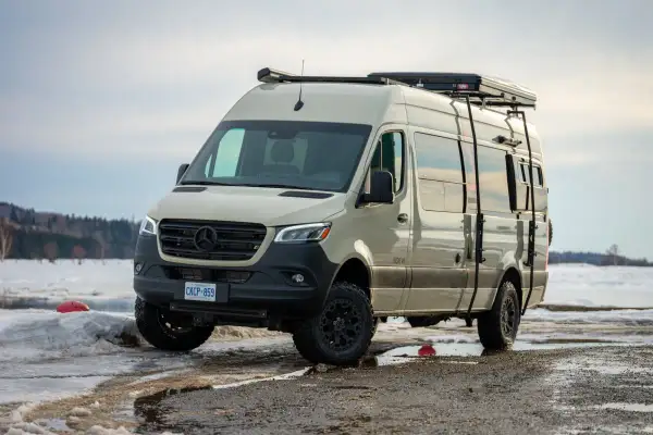 Una van 4x4 color arena, lista para la aventura invernal, con equipo de campamento en el techo y escaleras laterales, aparcada en un paisaje nevado.