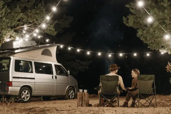 Pareja acampando bajo luces de feria en la costa atlántica francesa. Una furgoneta camper y sillas plegables crean un ambiente acogedor.