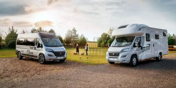 Dos vehículos, un camper y una caravana, estacionados al aire libre. Dos mujeres y sus perros disfrutan de un día soleado.